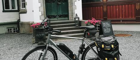Un vélo d’appartement équipé d’un panier et d’un petit sac, garé sur une surface de gravier devant un bâtiment avec une porte verte et une enseigne.