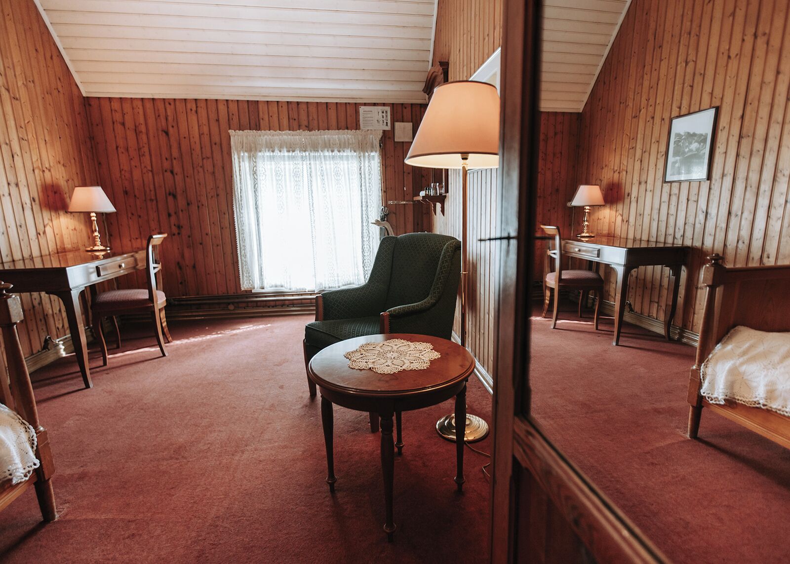 A room with wooden paneling, a green armchair, a round table with a lace tablecloth, and two bedside tables with lamps.