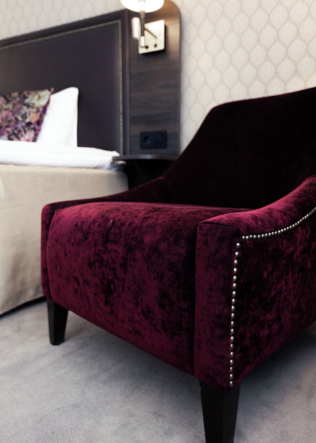 A dark red velvet armchair with a high back and decorative studs, positioned in a hotel room with a bed and nightstand in the background.