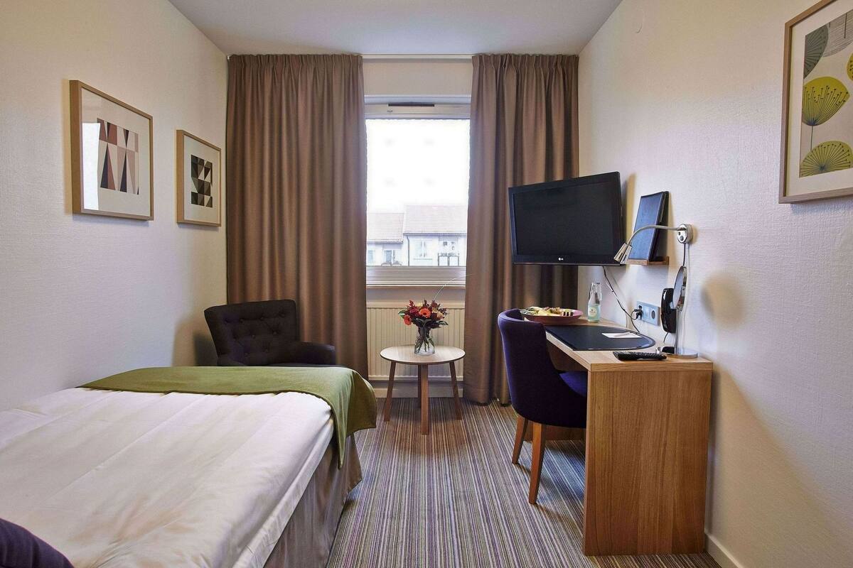 A hotel room with a bed, a desk with a computer, a chair, a TV, and a window with curtains.