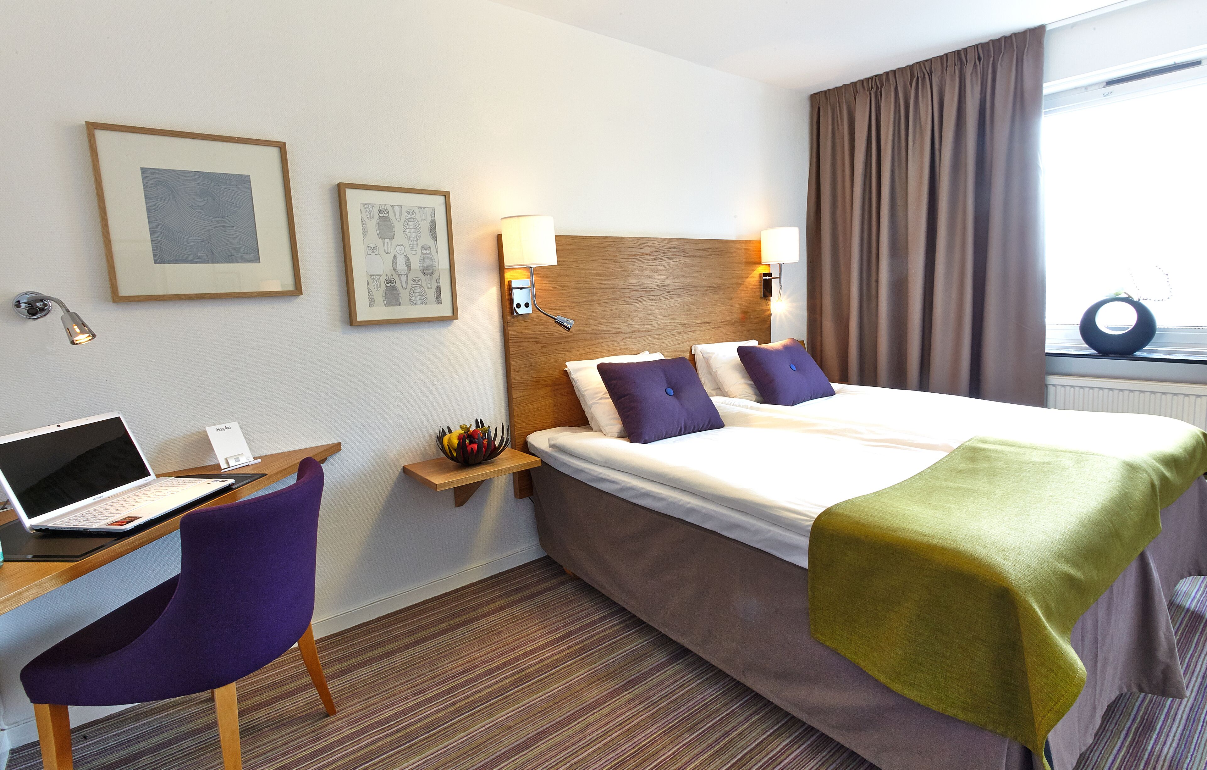 A hotel room with a bed, a desk with a laptop, a chair, two wall-mounted lamps, and framed artwork on the wall.