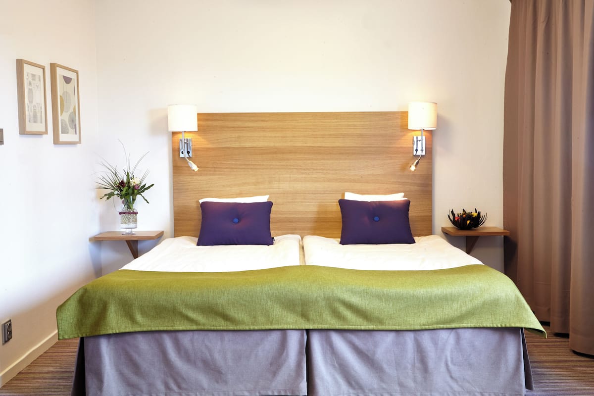A hotel room with a wooden headboard, two bedside tables with lamps, a green bedspread, and a small plant on one table.