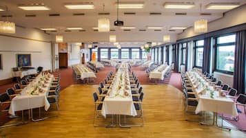 Un amplio salón de conferencias con filas de mesas y sillas, ventanas grandes y candelabros.