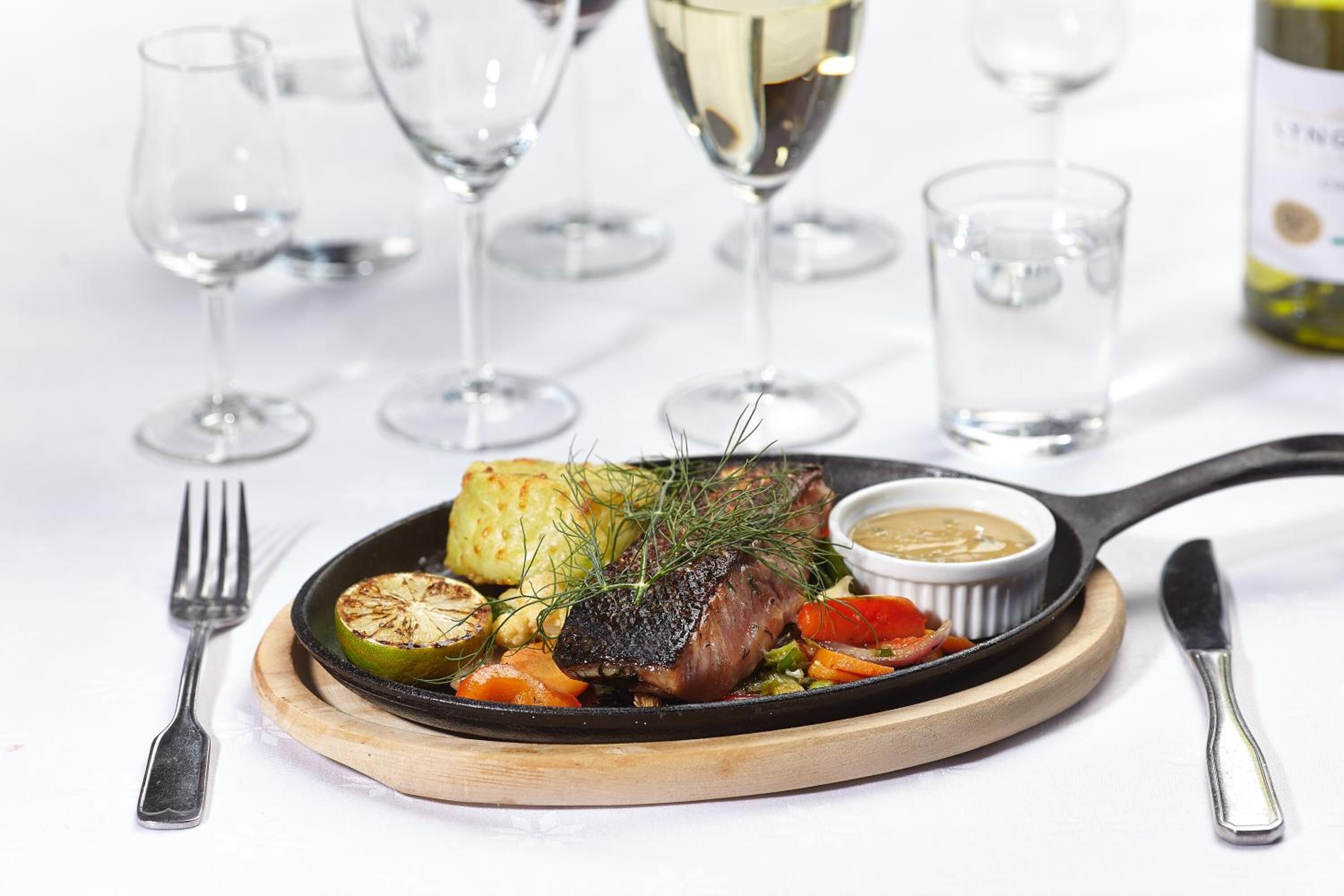 A plate of food with a steak, vegetables, and a sauce, accompanied by a fork and knife on a wooden board.