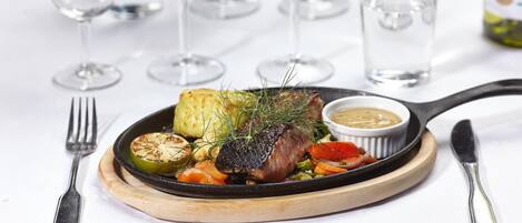 A plate of food with a steak, vegetables, and a sauce, accompanied by a fork and knife on a wooden board.