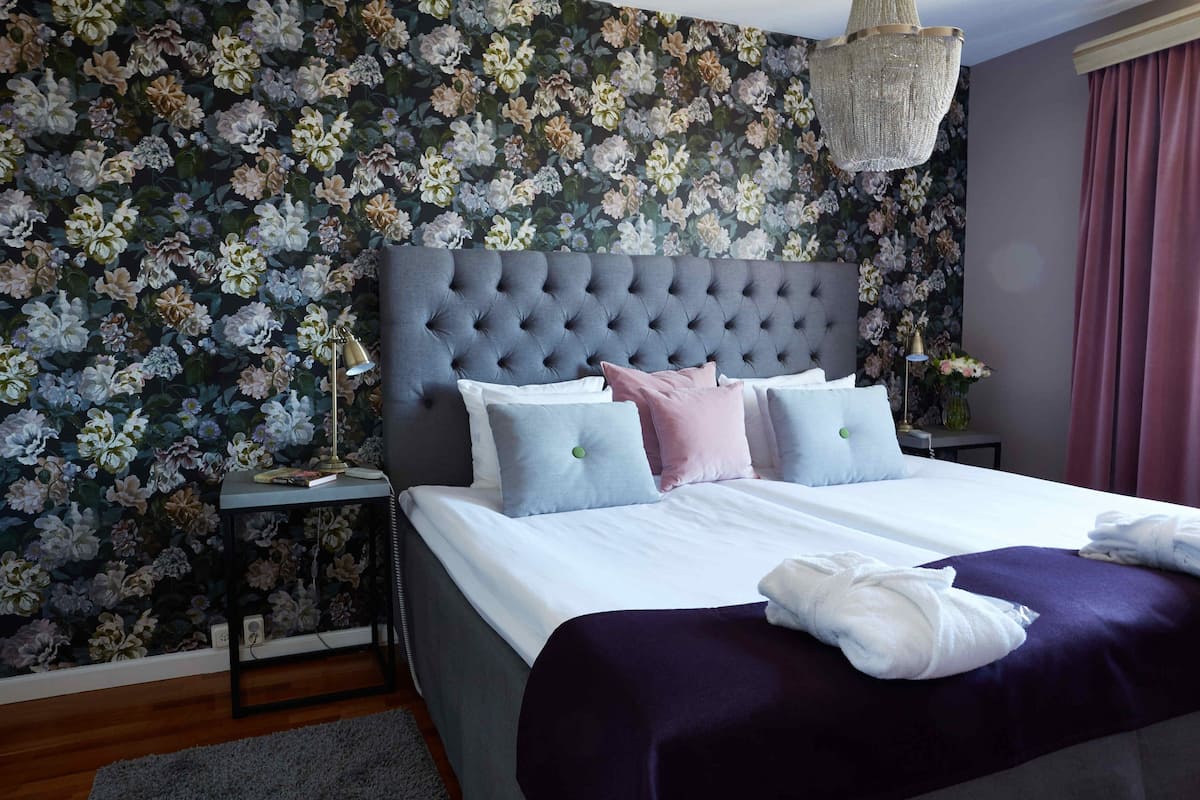 A neatly made bed with a tufted headboard, a bedside table with a lamp, and a floral wallpaper backdrop.