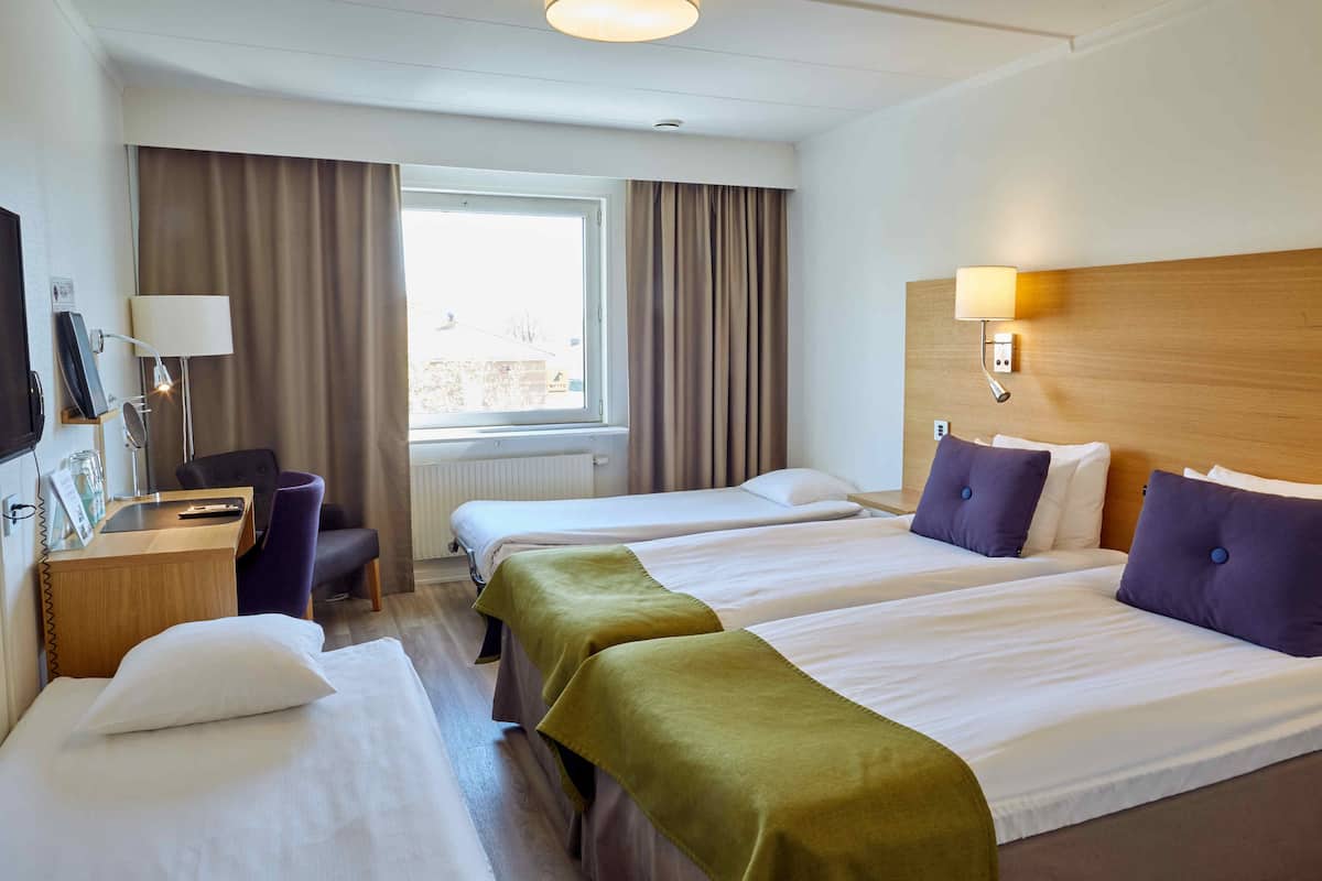 A hotel room with two beds, a desk, a chair, a lamp, and a window with curtains.