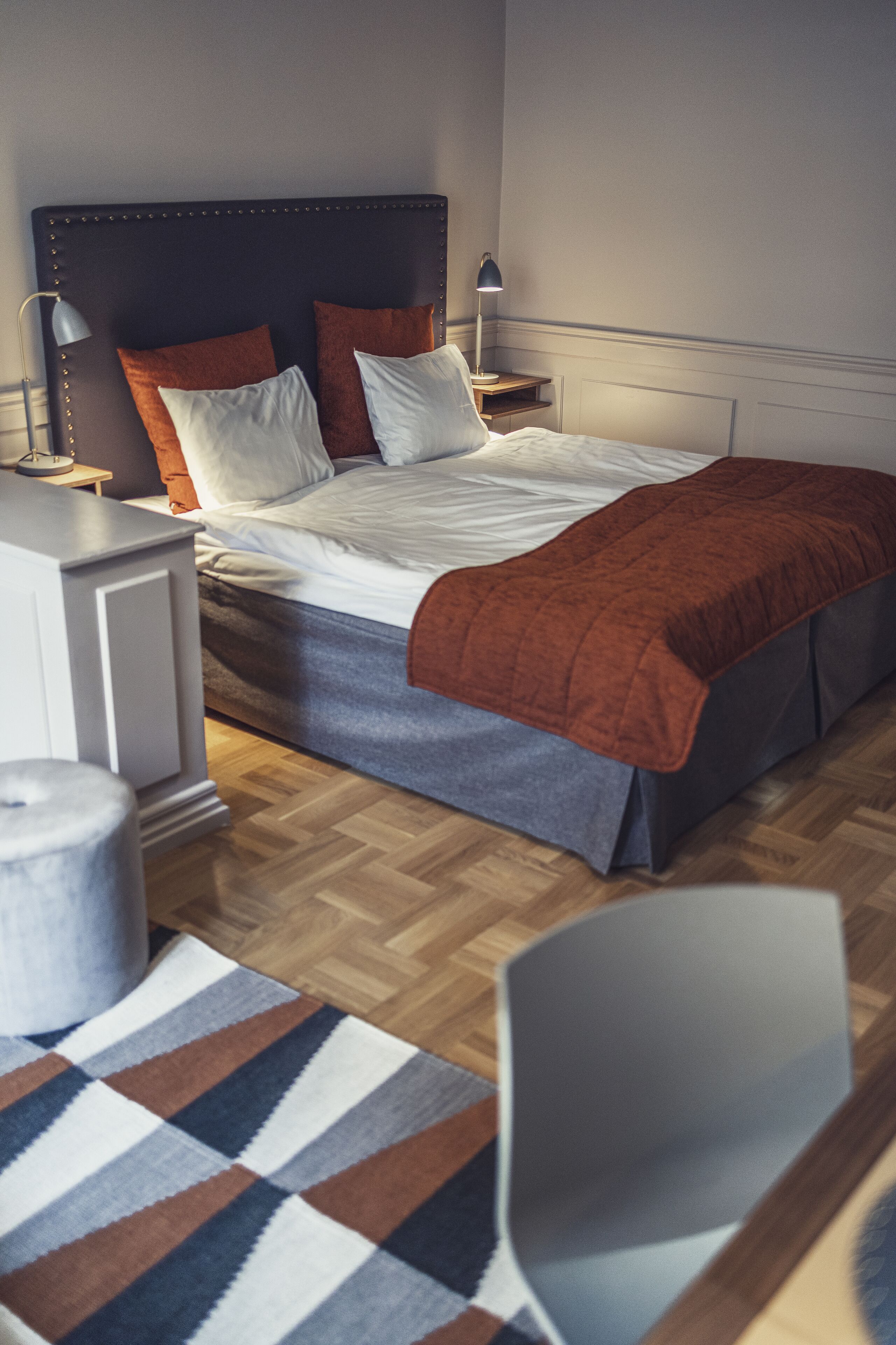 Un dormitorio ordenado con cama, mesitas de noche, lámpara, silla y alfombra estampada.
