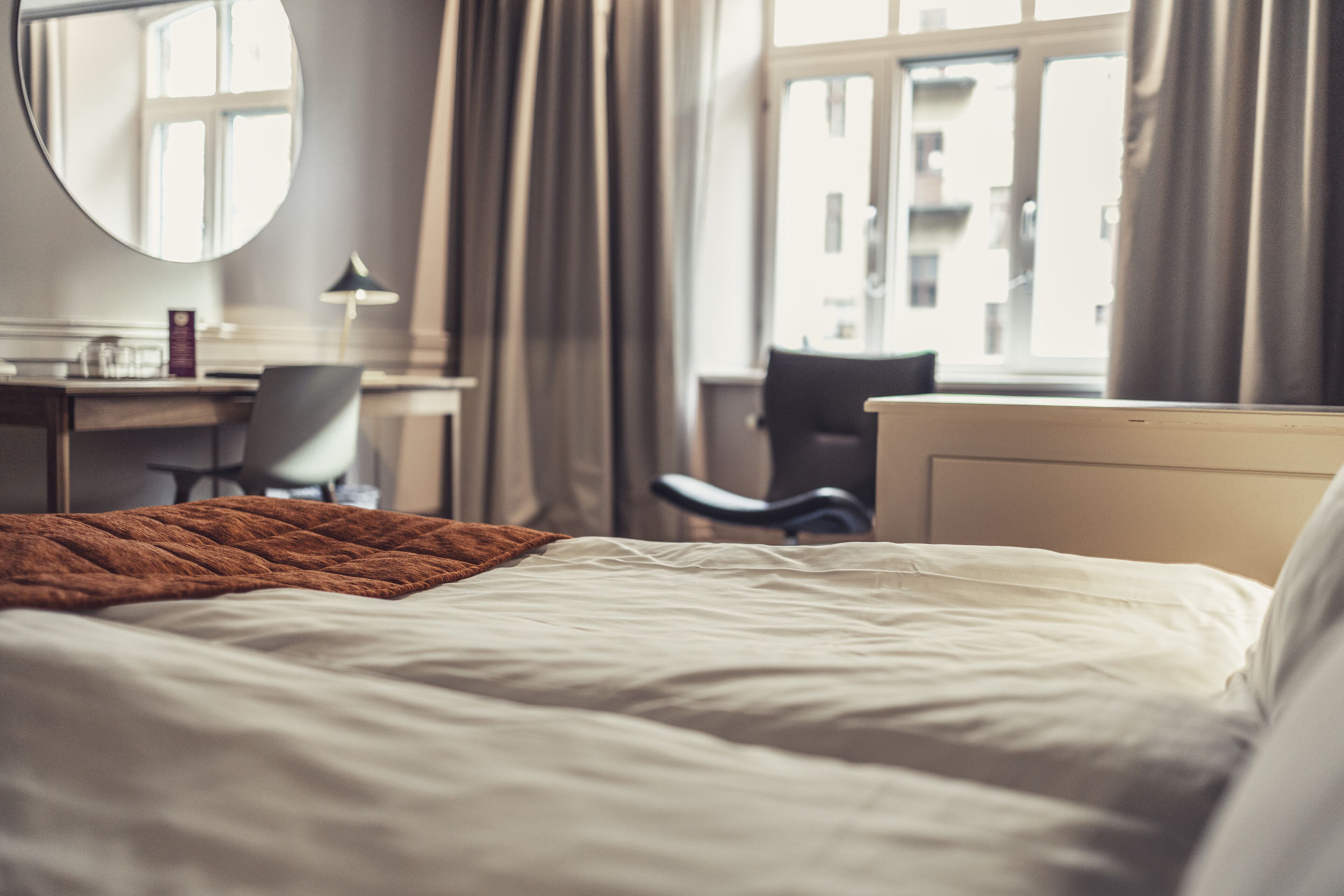 Ett hotellrum med en säng, ett skrivbord, en stol, en rund spegel och ett fönster med gardiner.
