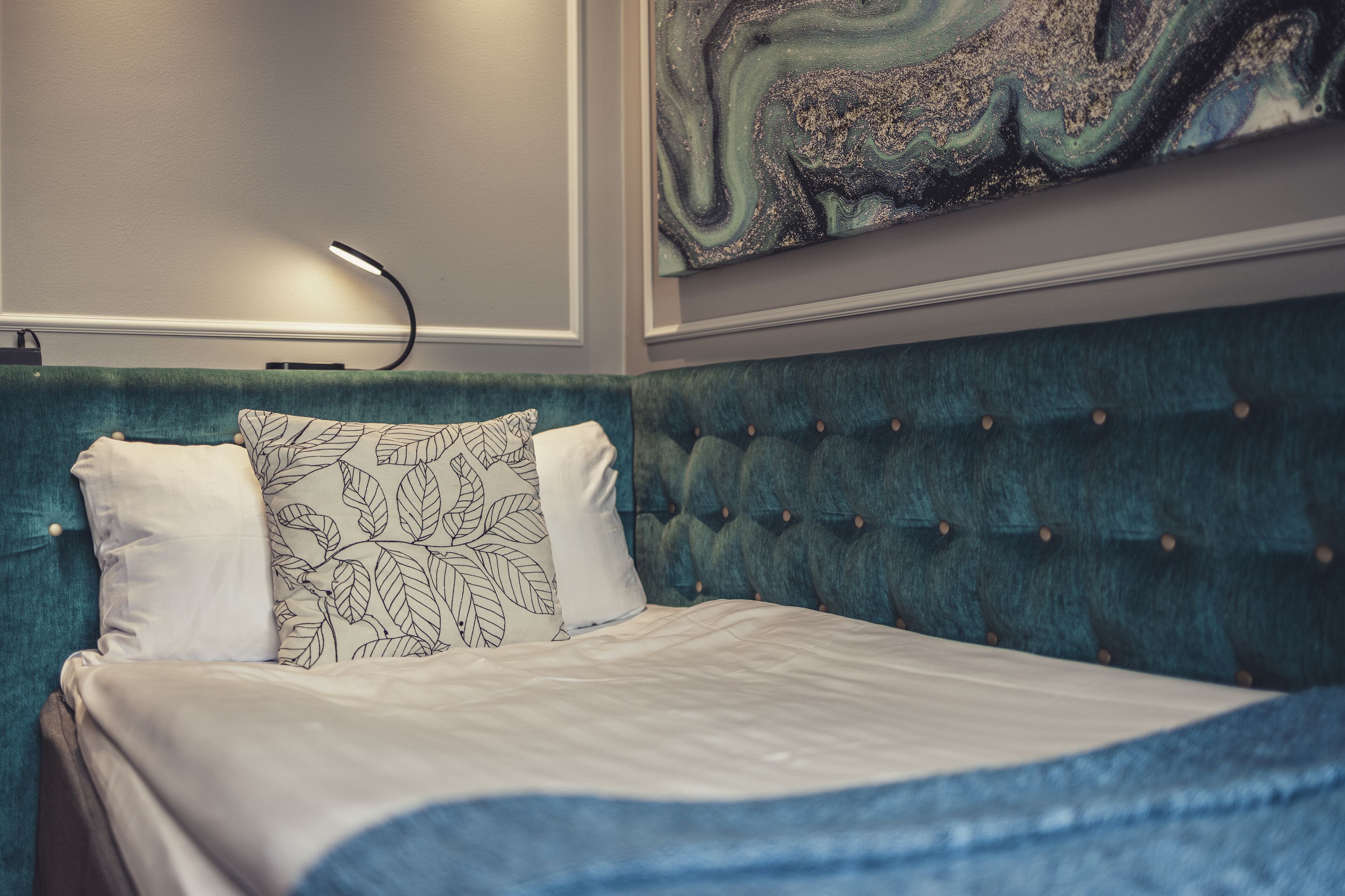 Una cama con cabecera color aguamarina, ropa de cama blanca y una almohada con estampado de hojas.