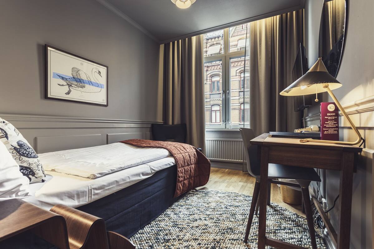 A hotel room with a bed, a desk, a chair, a framed picture on the wall, and a window with curtains.