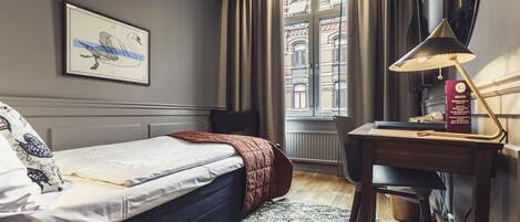 Habitación de hotel con cama, escritorio, silla, un cuadro en la pared y ventana con cortinas.