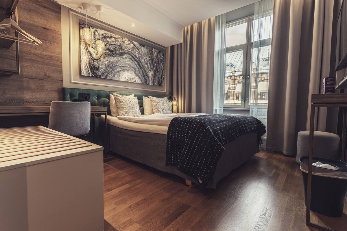 A modern hotel room with a large bed, a wooden desk, a green upholstered headboard, and a large abstract painting on the wall.