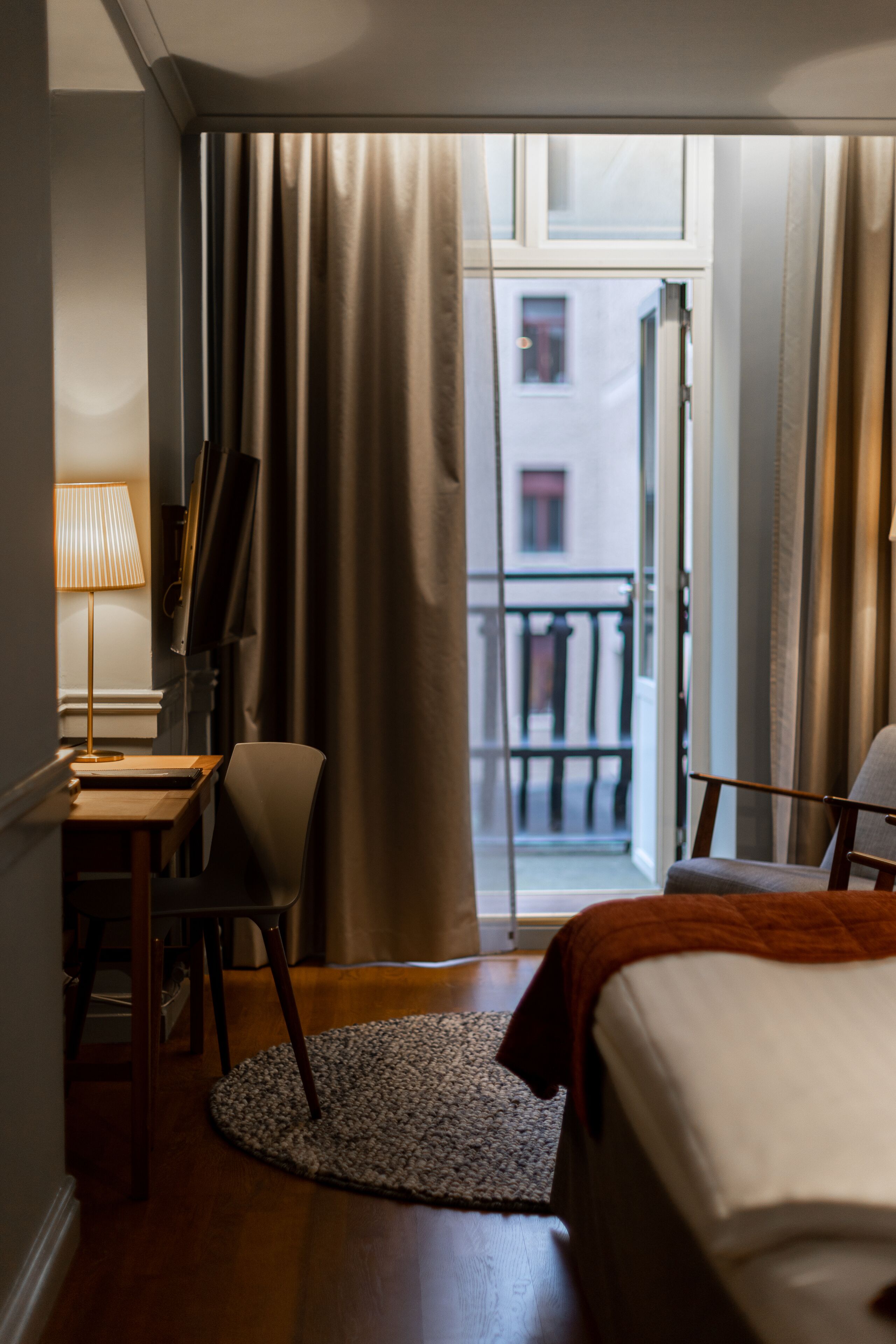 A hotel room with a bed, a desk with a lamp, a chair, a small rug, and a view of the outside balcony.