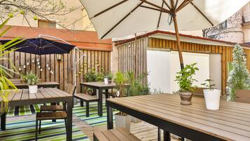 An outdoor patio area with wooden tables and benches, potted plants, and a large umbrella providing shade.