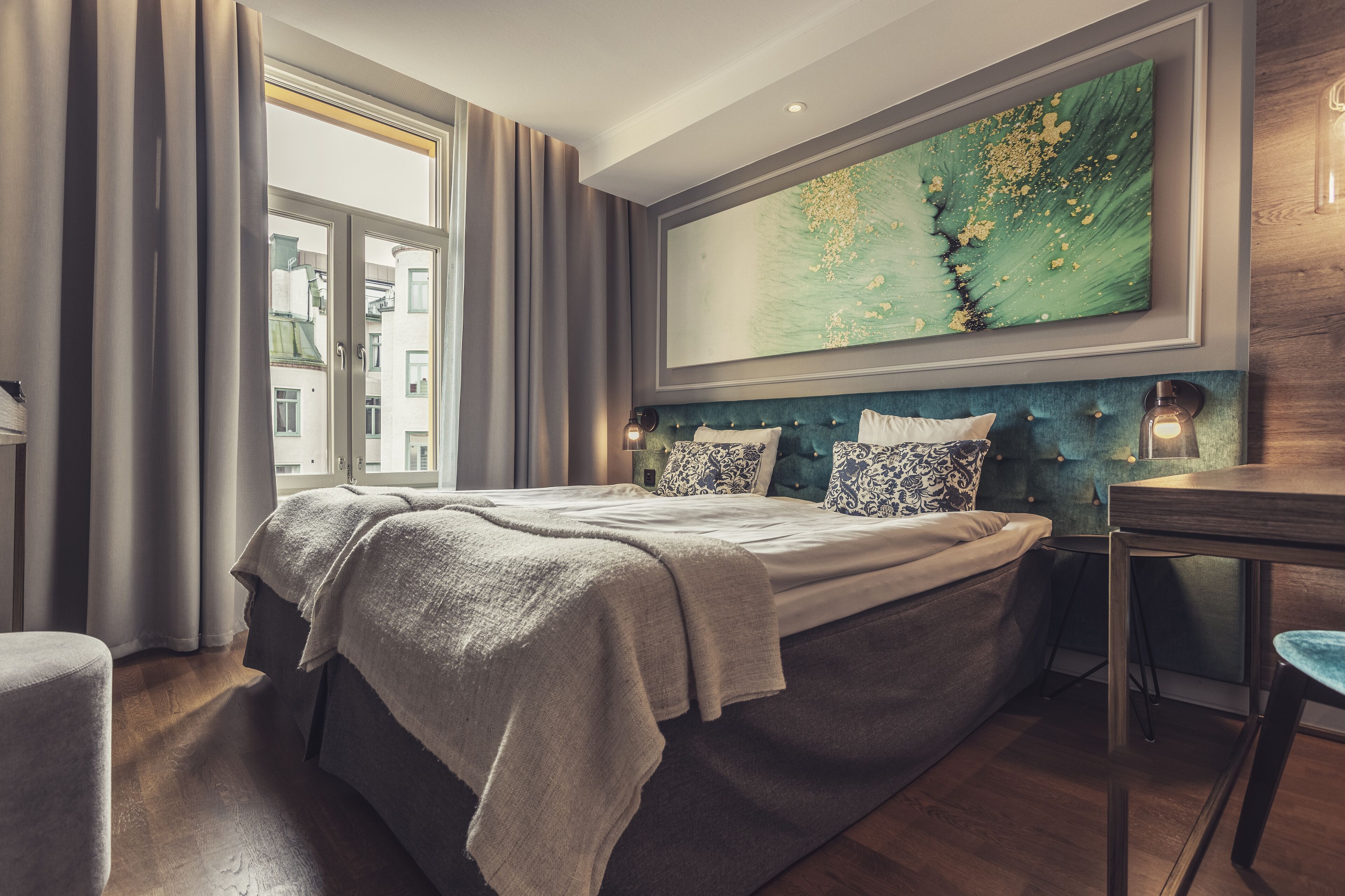 A modern bedroom with a large bed, a headboard with built-in lighting, a view of the city through a window, and a large abstract painting above the bed.