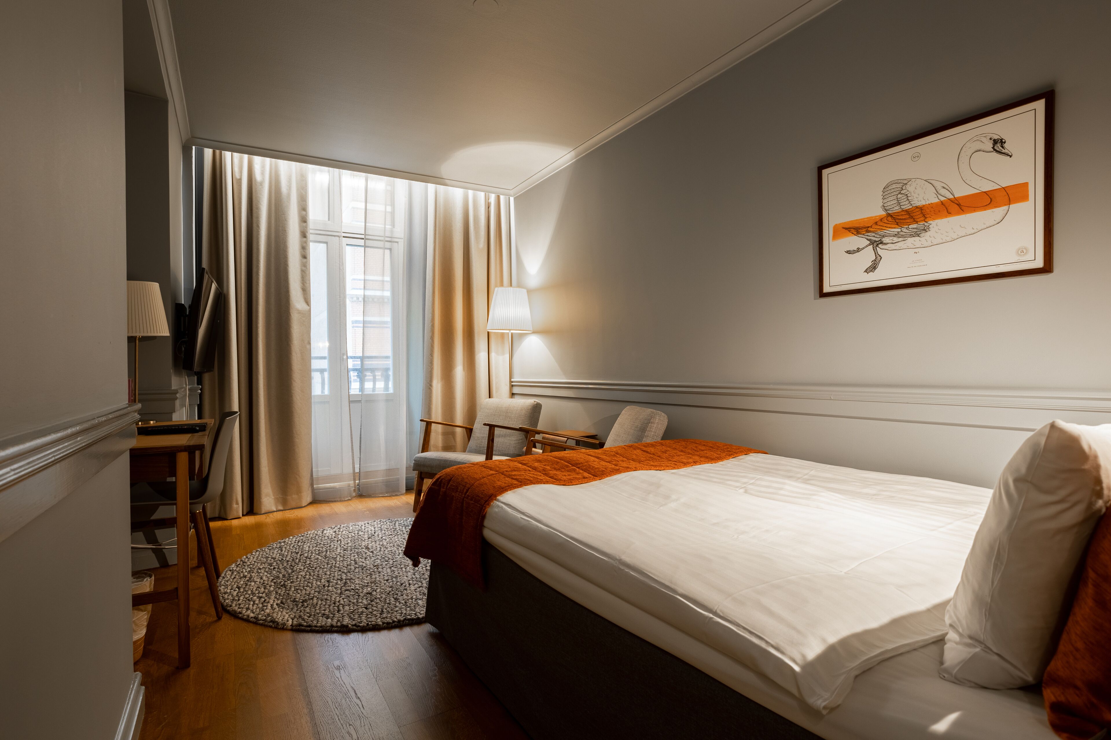 Una habitación de hotel con cama, escritorio, silla, lámpara y ventana con cortinas.