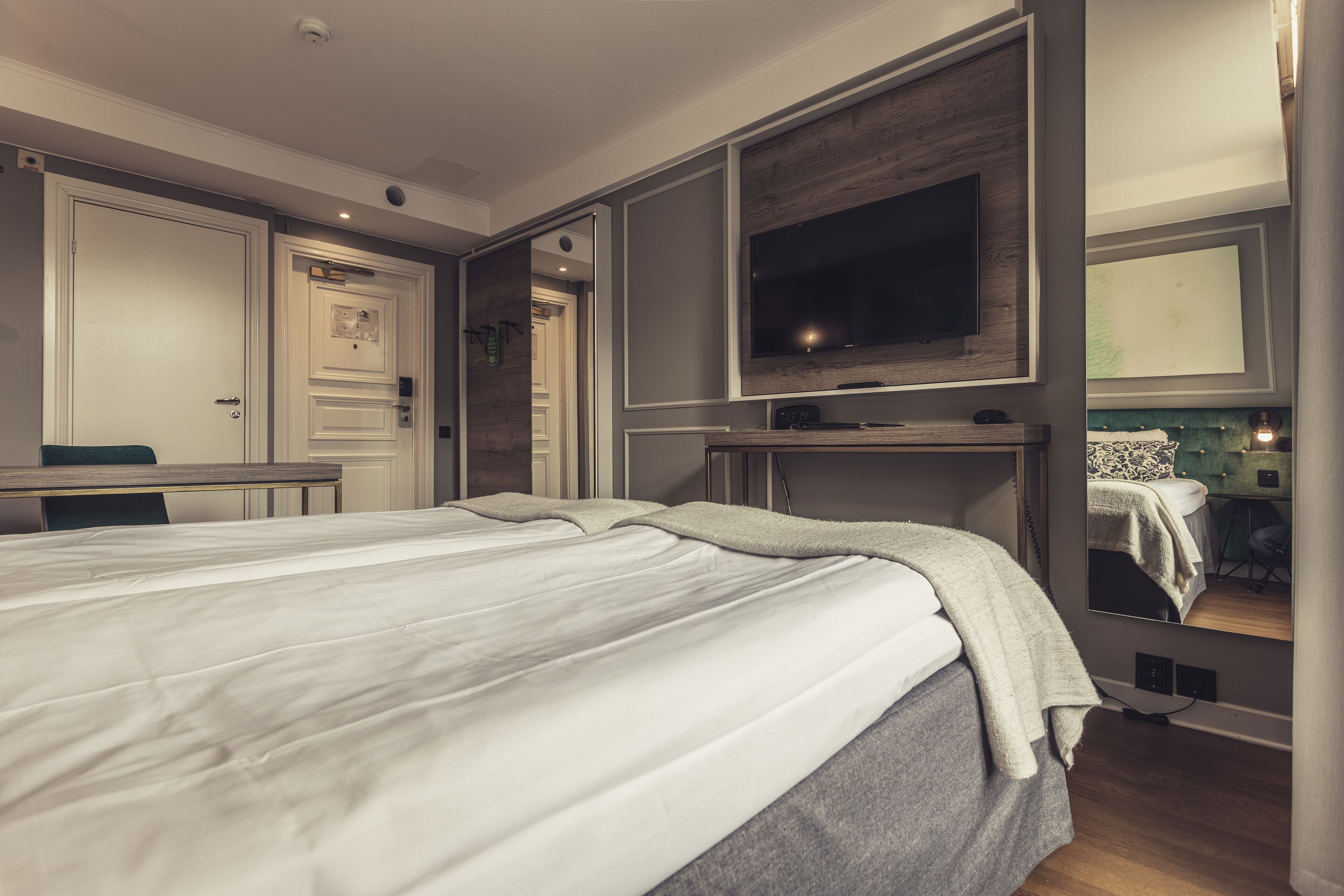 A modern hotel room with a large bed, a desk, a TV mounted on the wall, and a mirror reflecting the room.