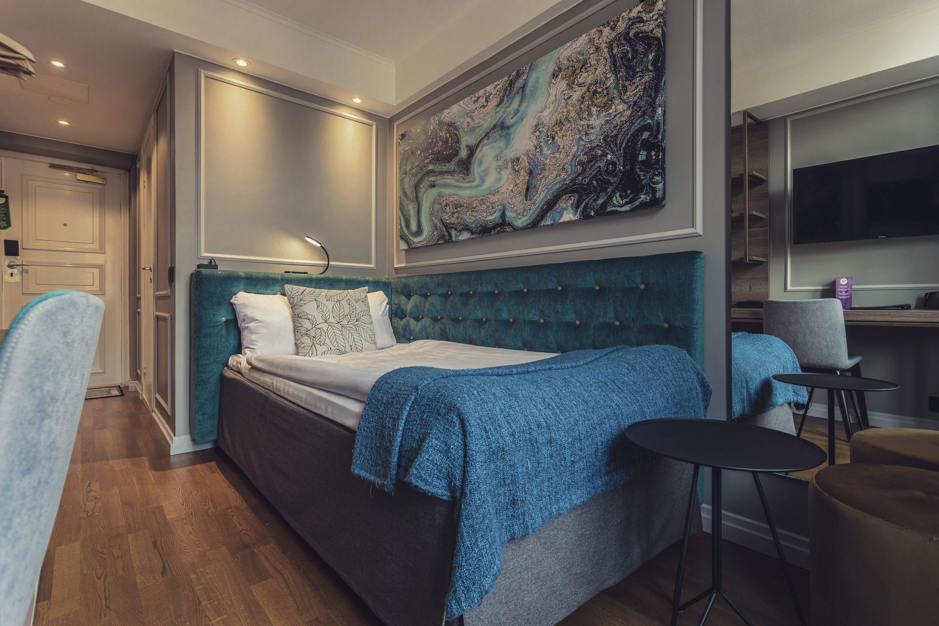Un dormitorio con una cama grande, una colcha azul, un cabecero moderno, un cuadro en la pared y un televisor a la derecha.