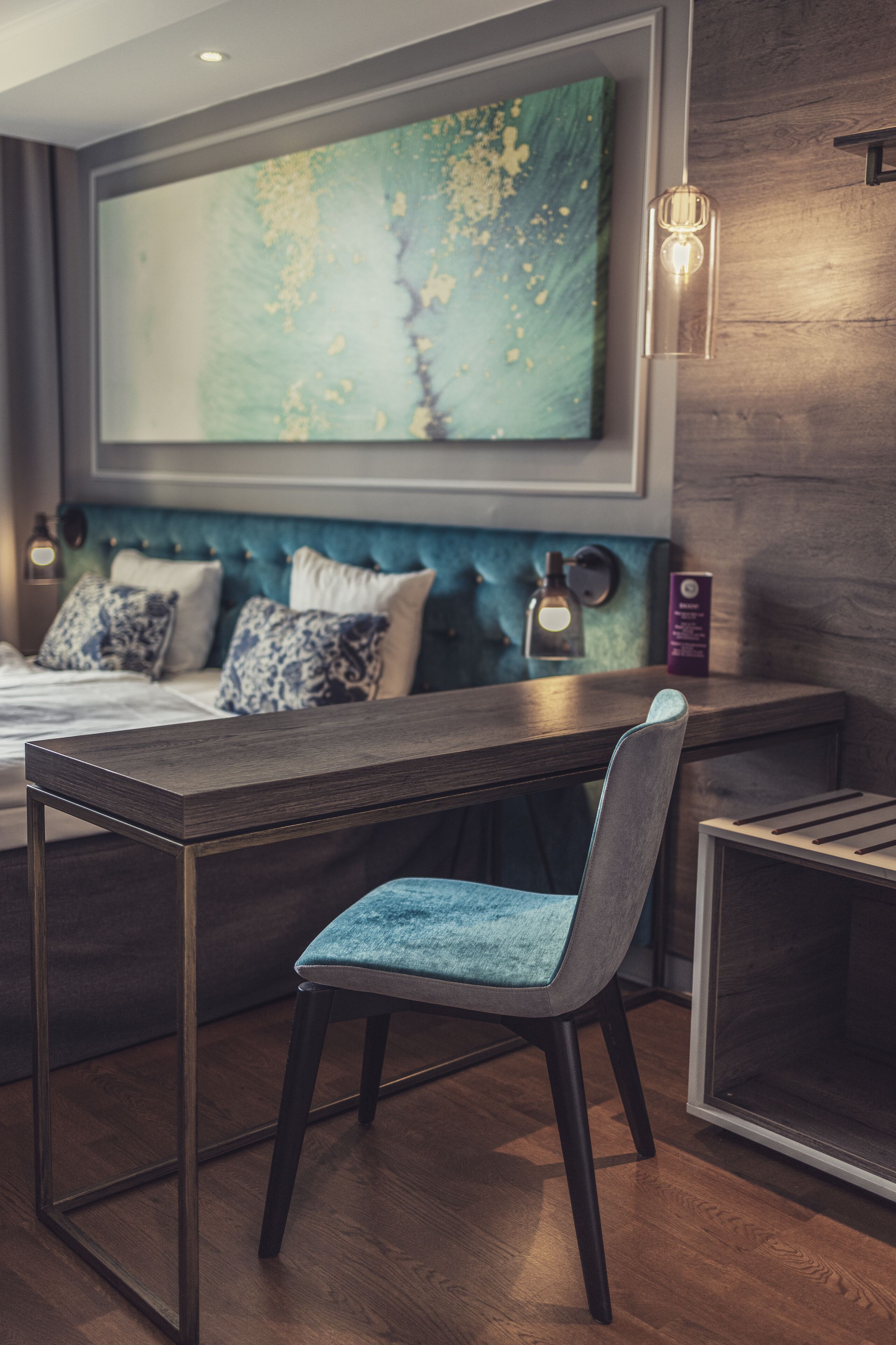 A modern bedroom with a bed, a desk, and a chair. There is a large abstract painting on the wall and a small table with a lamp.