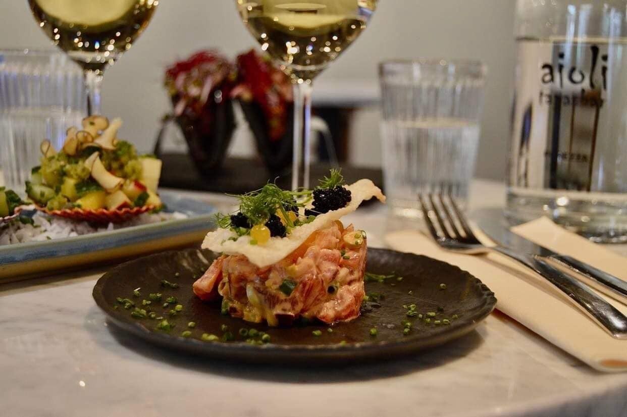 Una mesa de comedor con un plato de comida, copas de vino blanco y una botella de agua.