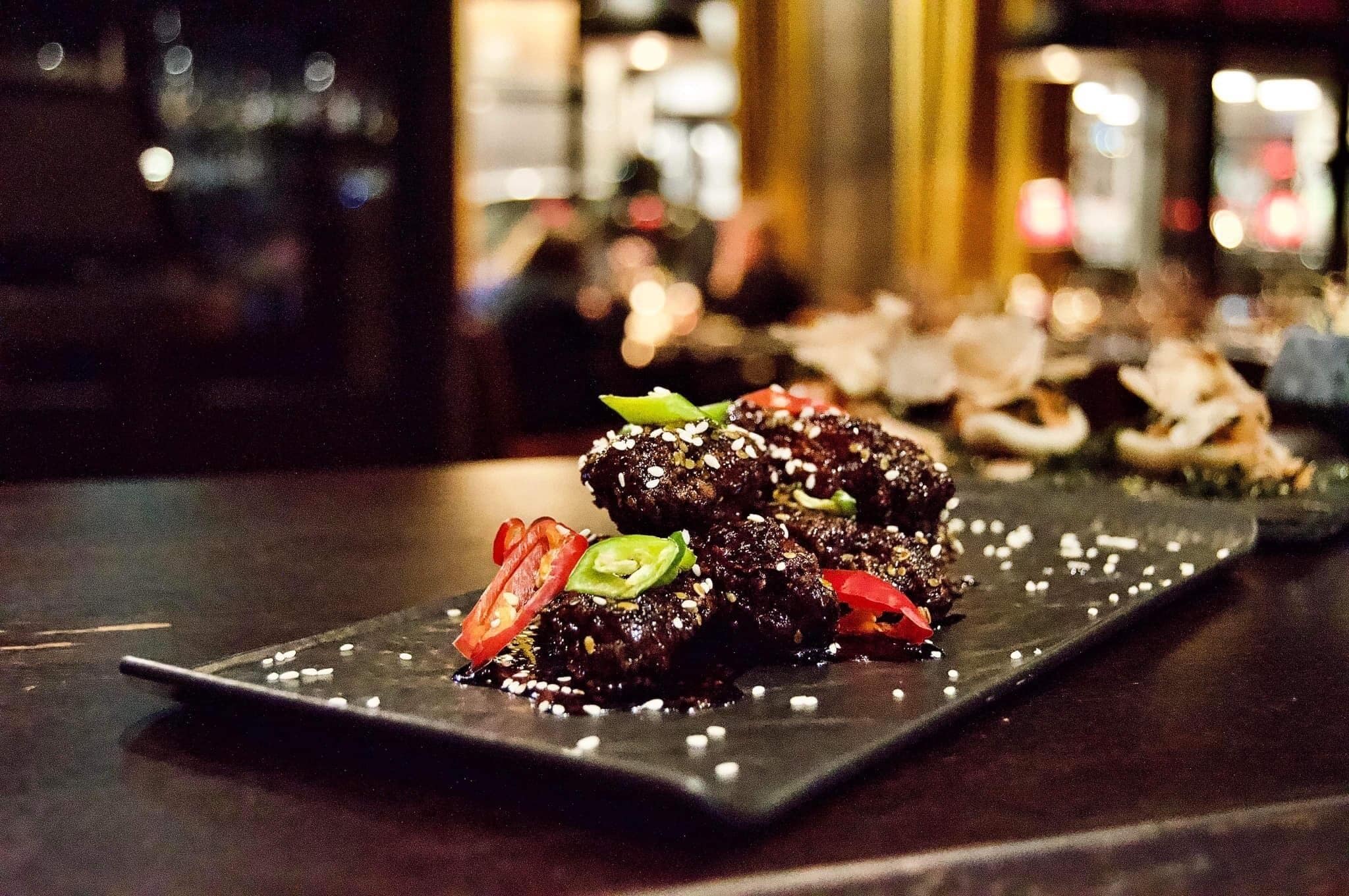 Un plat de boulettes de viande laquées, garnies de graines de sésame et de légumes coupés en tranches, servi dans une assiette foncée.