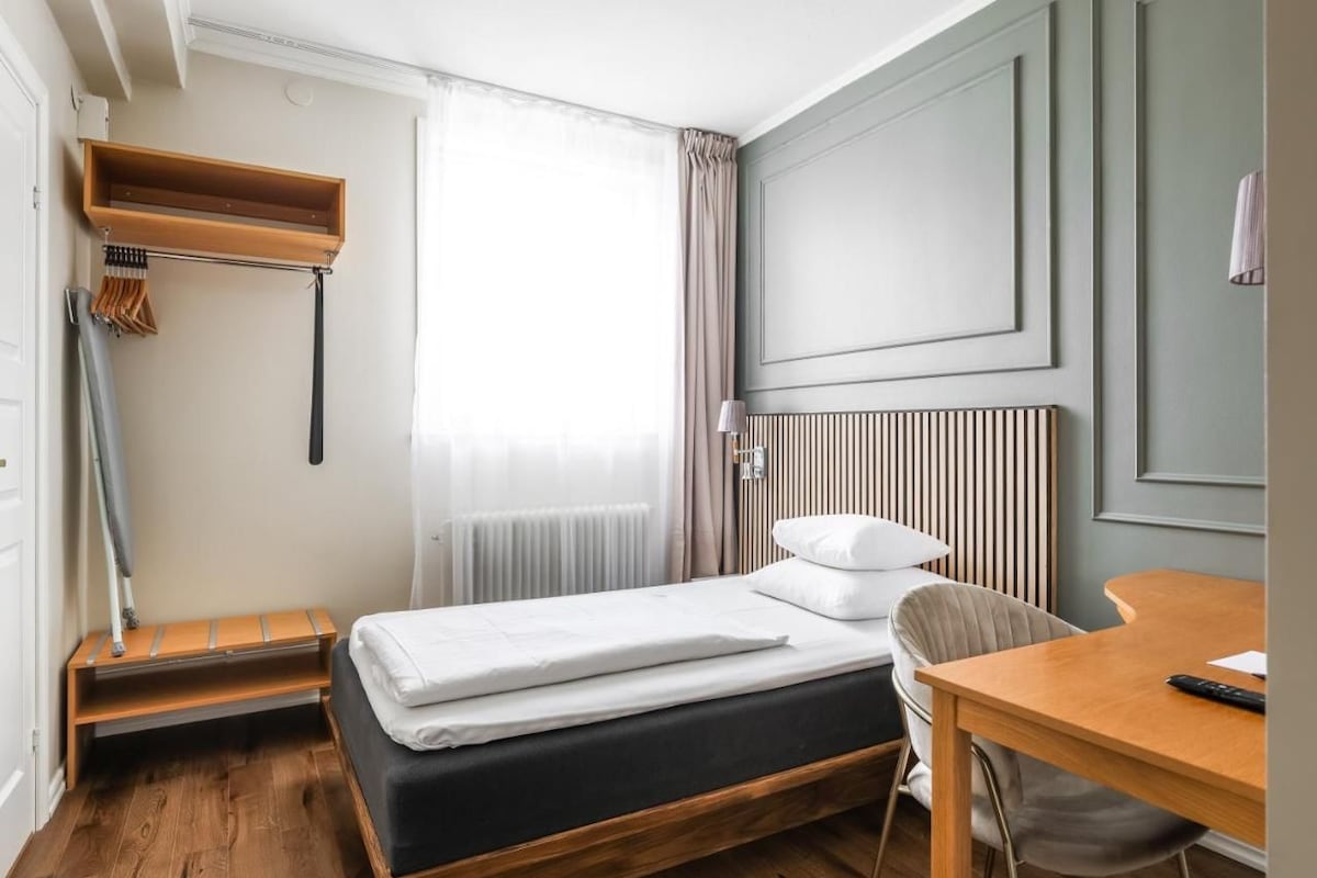 A compact hotel room with a single bed, a small table, a chair, a radiator, and a window with curtains.