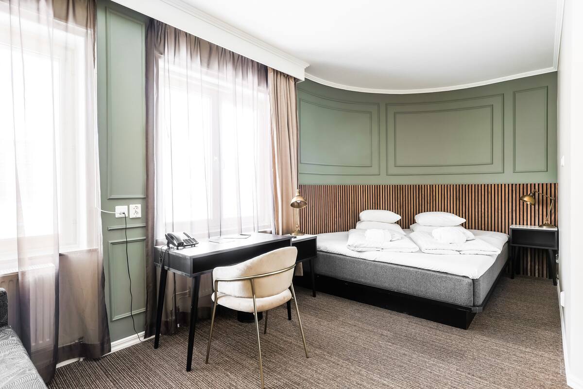 A hotel room with a bed, desk, chair, and a window with sheer curtains.