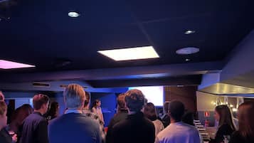 Un salón de conferencias con asistentes mirando una pantalla de presentación, equipado con luces de techo y un extintor visible.