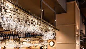 Zona de bar con una barra larga, copas de vino colgantes y una pared con diseño.