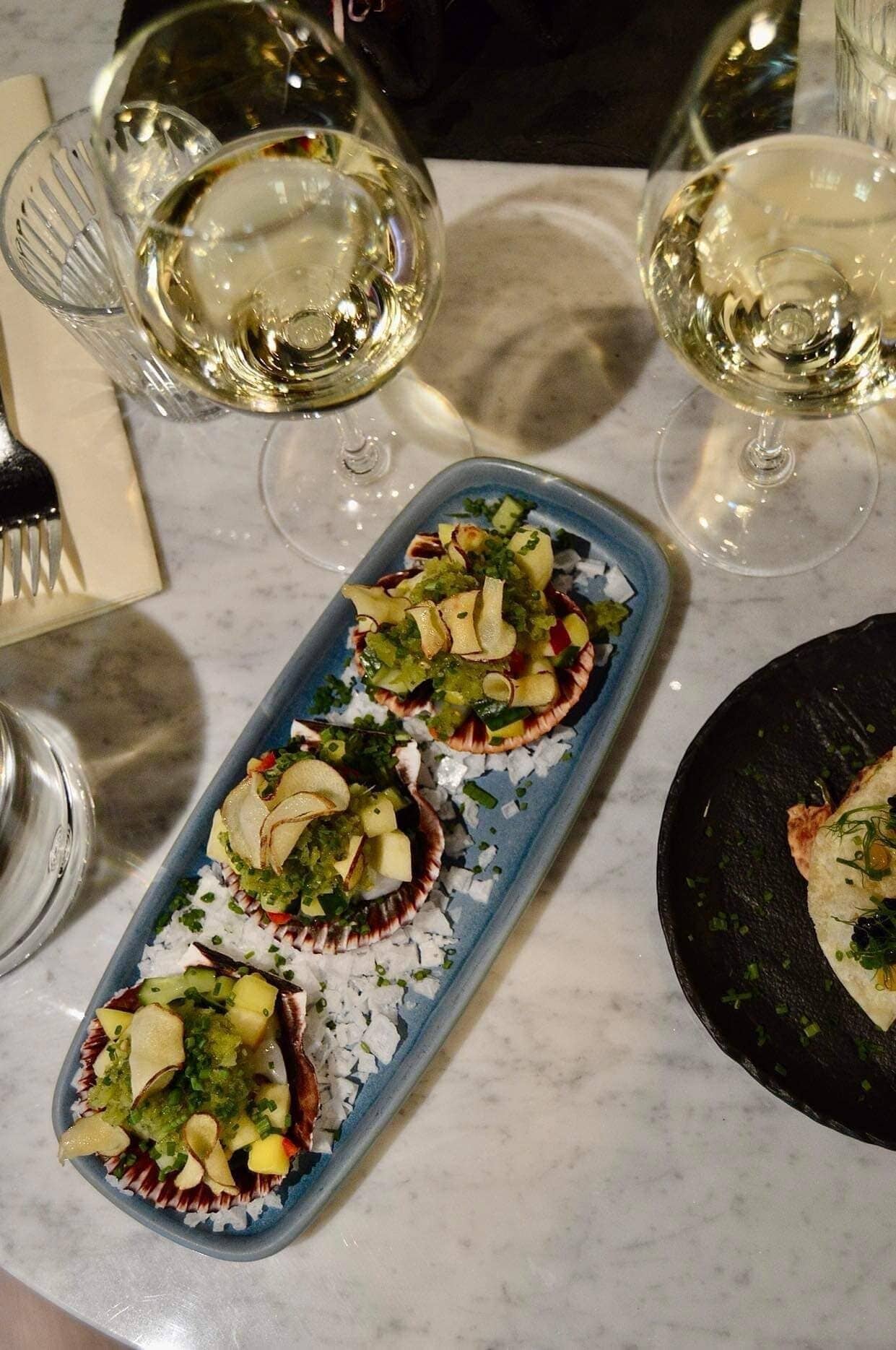 A dining table with a blue plate of scallops, white wine glasses, and a fork.