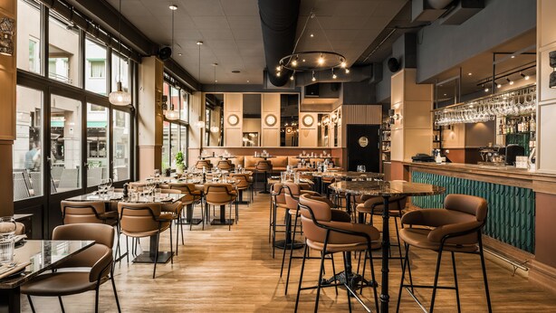 Un restaurante con varias mesas y sillas, una zona de bar y grandes ventanales que dejan entrar luz natural.