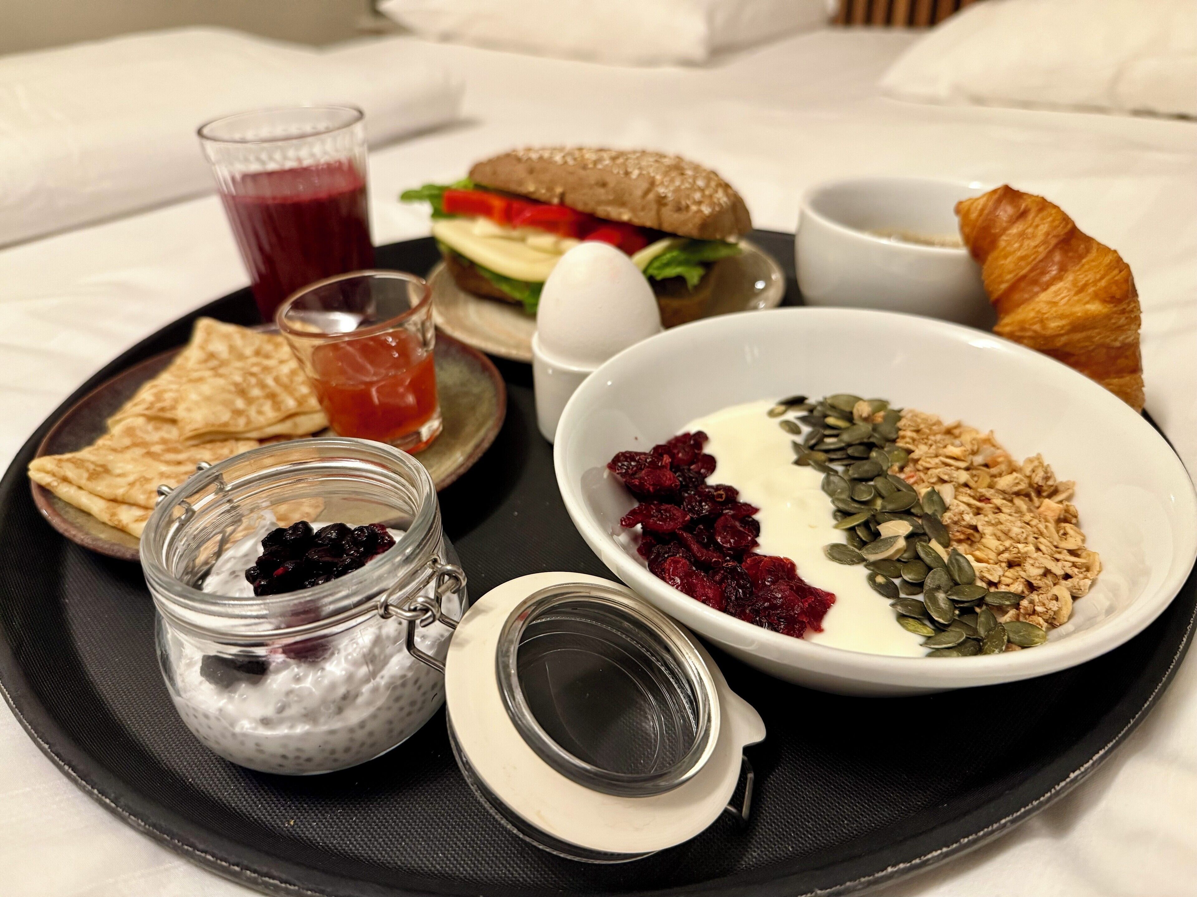 Un plateau de petit-déjeuner comprenant un croissant, un sandwich, un bol de yaourt aux graines, un verre de jus de fruits, un pot de fruits rouges et une assiette de toasts.