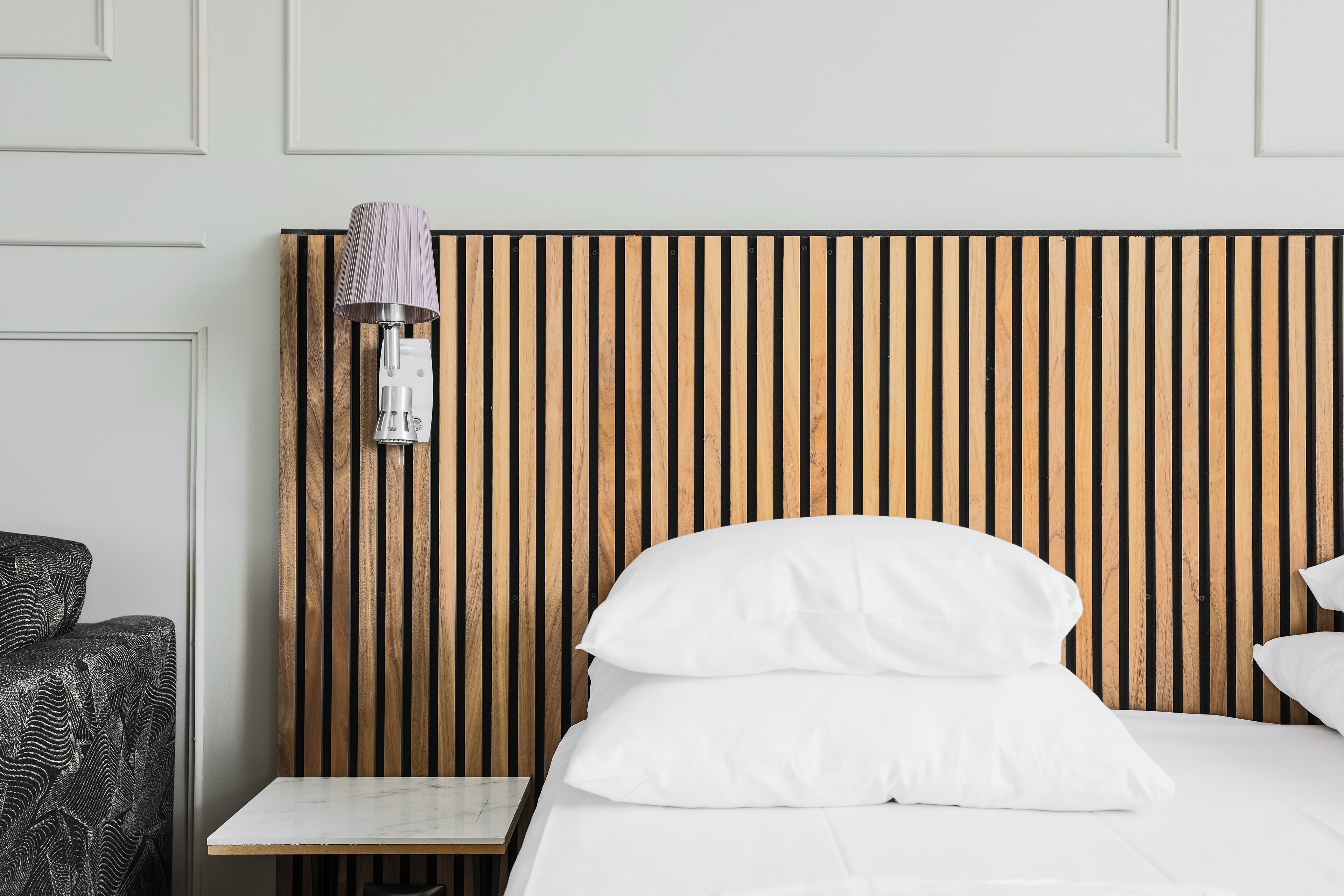 Una cama bien hecha con sábanas y almohadas blancas, una mesita de noche pequeña y una lámpara fijada a la pared.