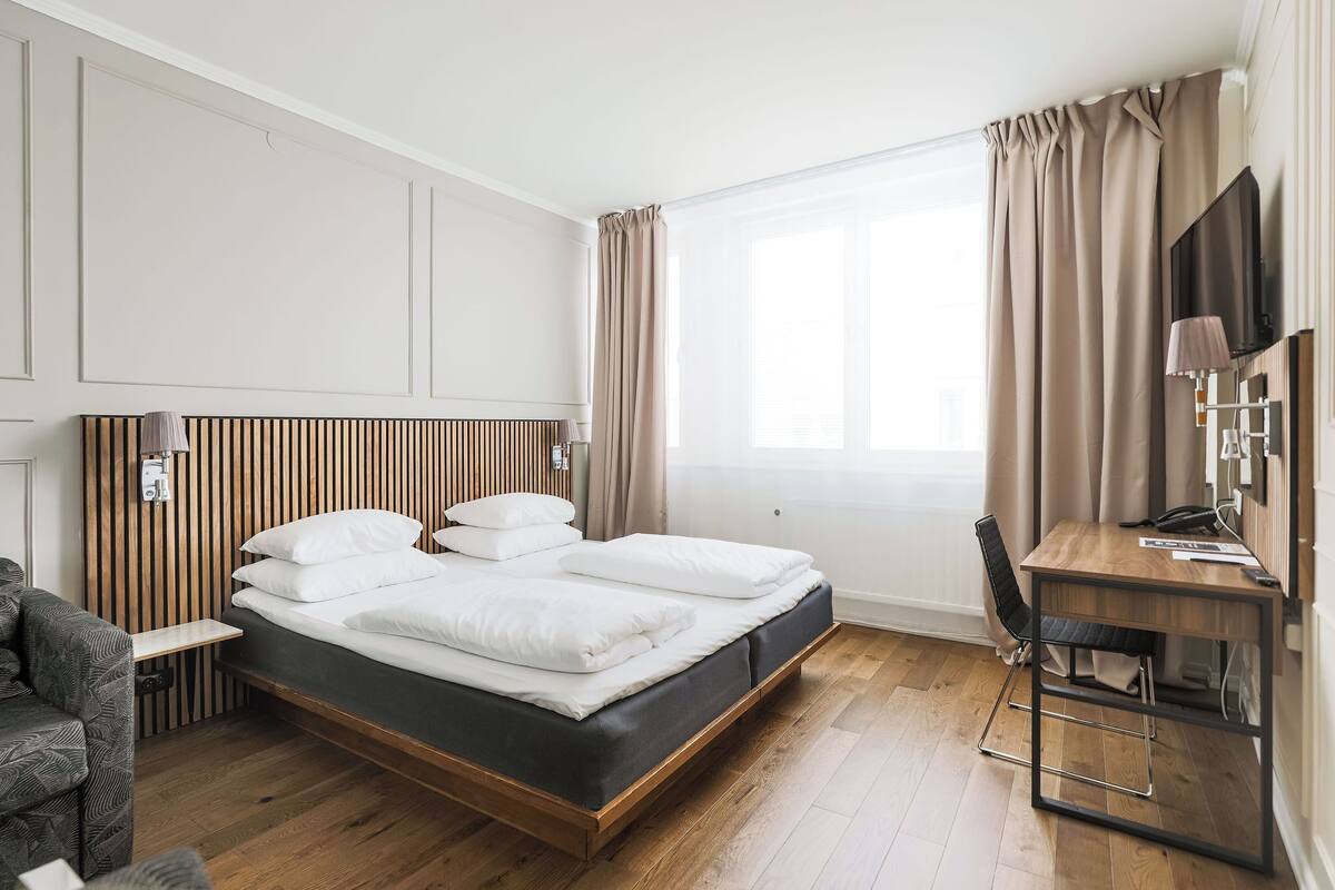 A hotel room with a wooden bed, a desk, a chair, and a large window with curtains.