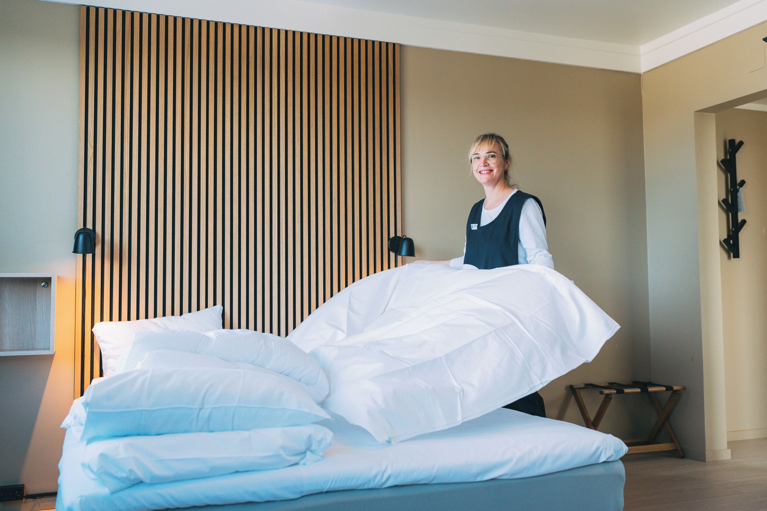 A neatly made bed with white linens in a modern room with a wooden slat wall and a small wooden stool.