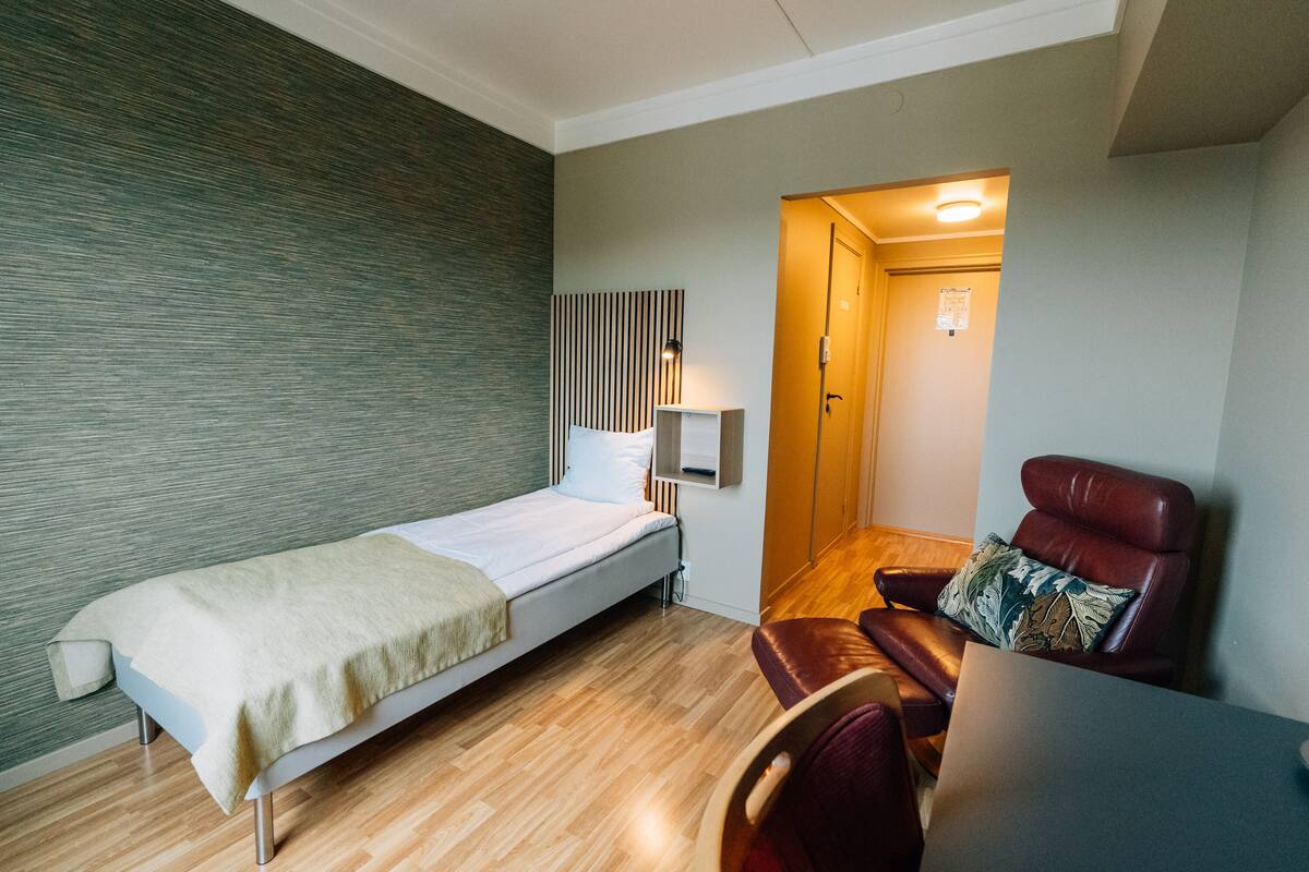 A single bed with a white mattress and a beige blanket, a leather armchair with a patterned pillow, a small table, and a wall-mounted lamp.