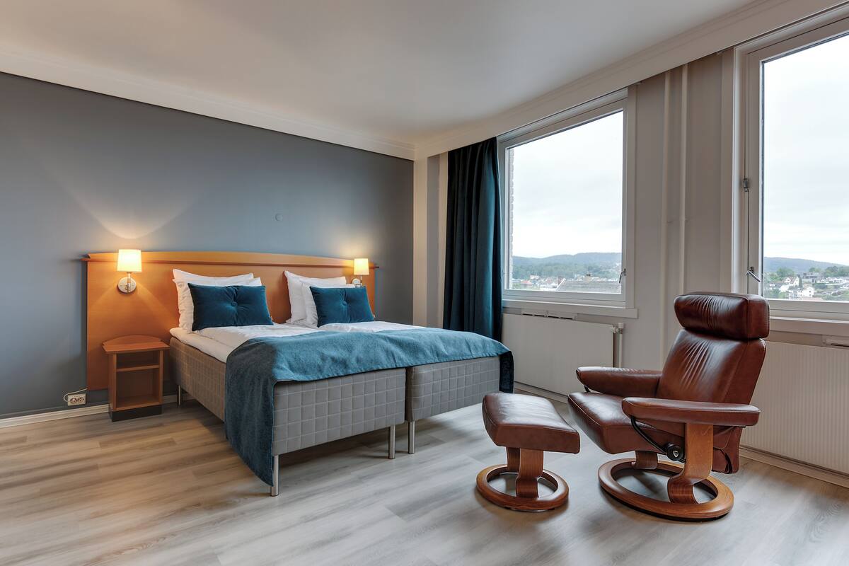 A modern hotel room with a bed, bedside table, chair, and large windows offering a city view.