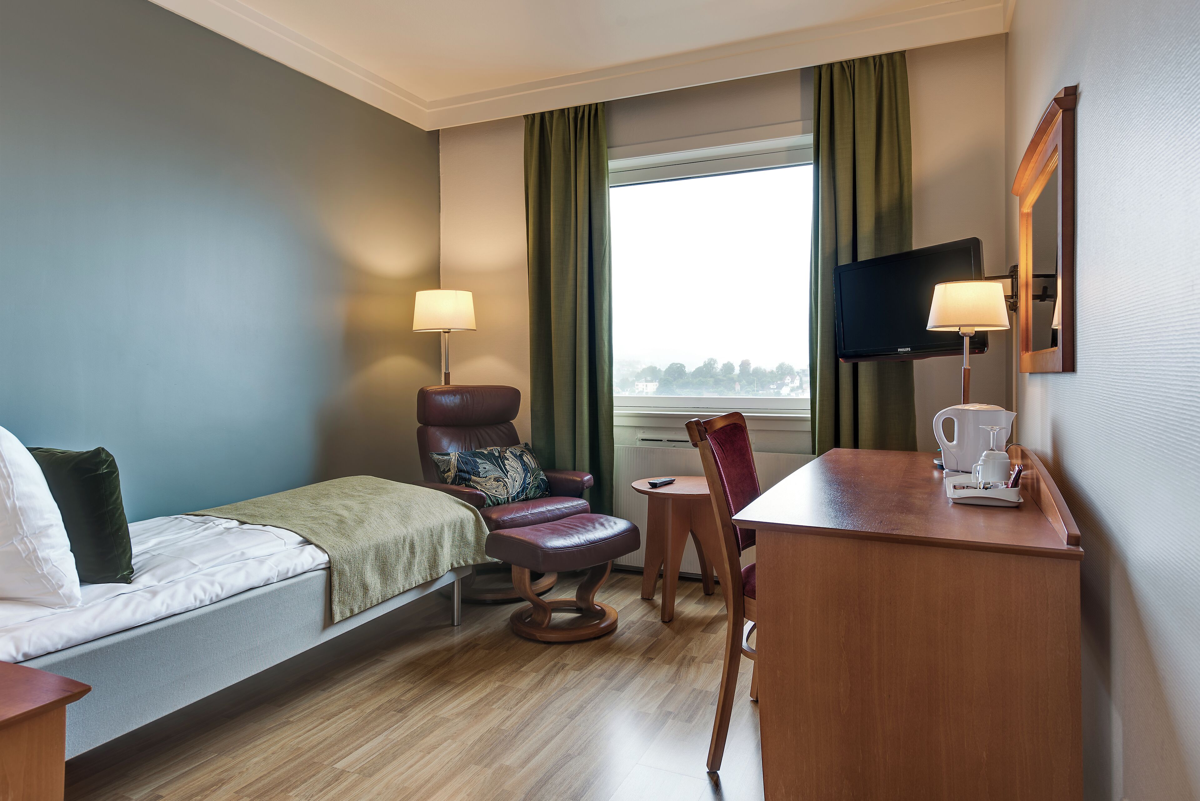 Una habitación de hotel con cama, una silla, un escritorio con cafetera, televisor y vista al exterior.