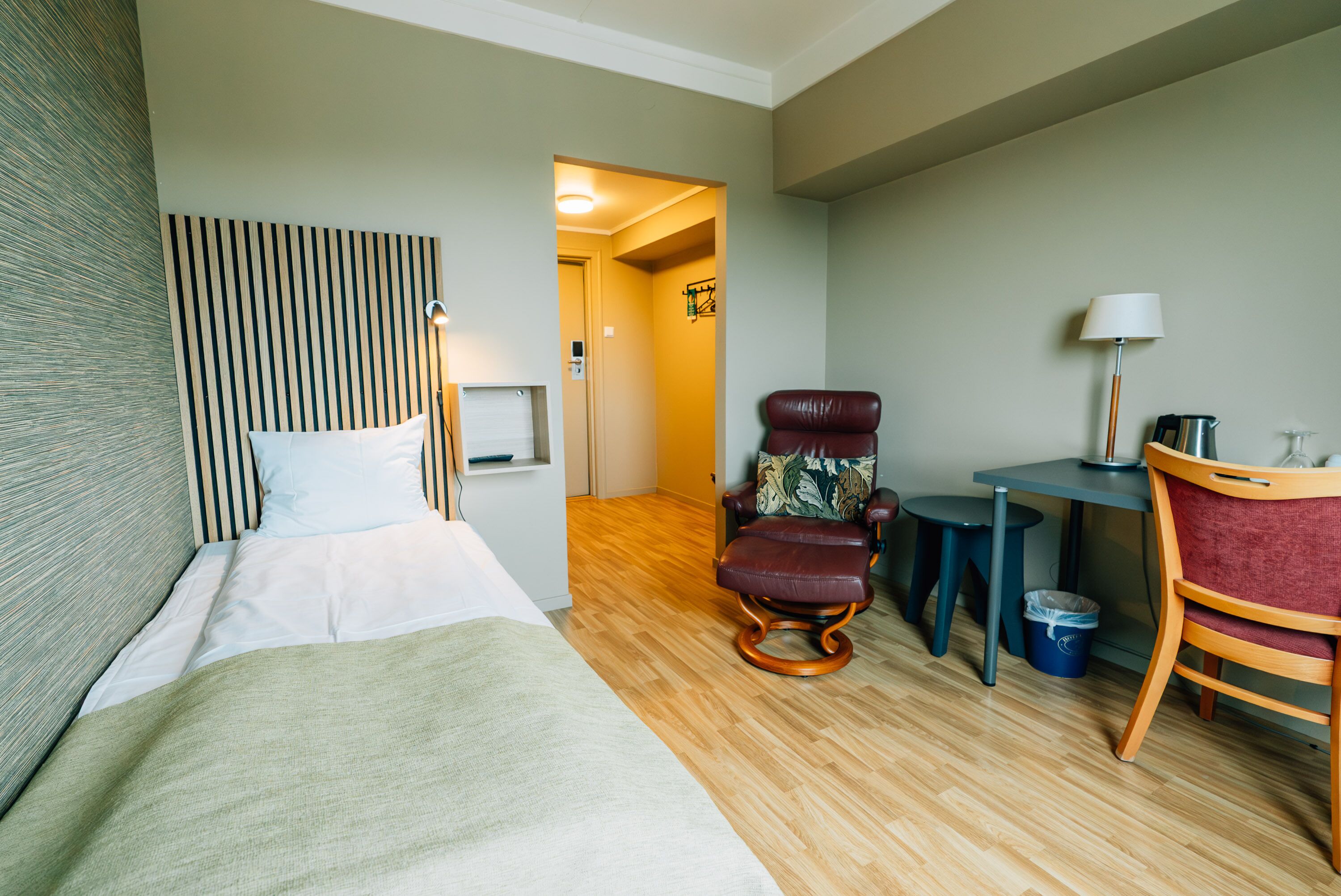 Ett hotellrum med en säng, en gungstol, ett skrivbord med en lampa och en papperskorg.