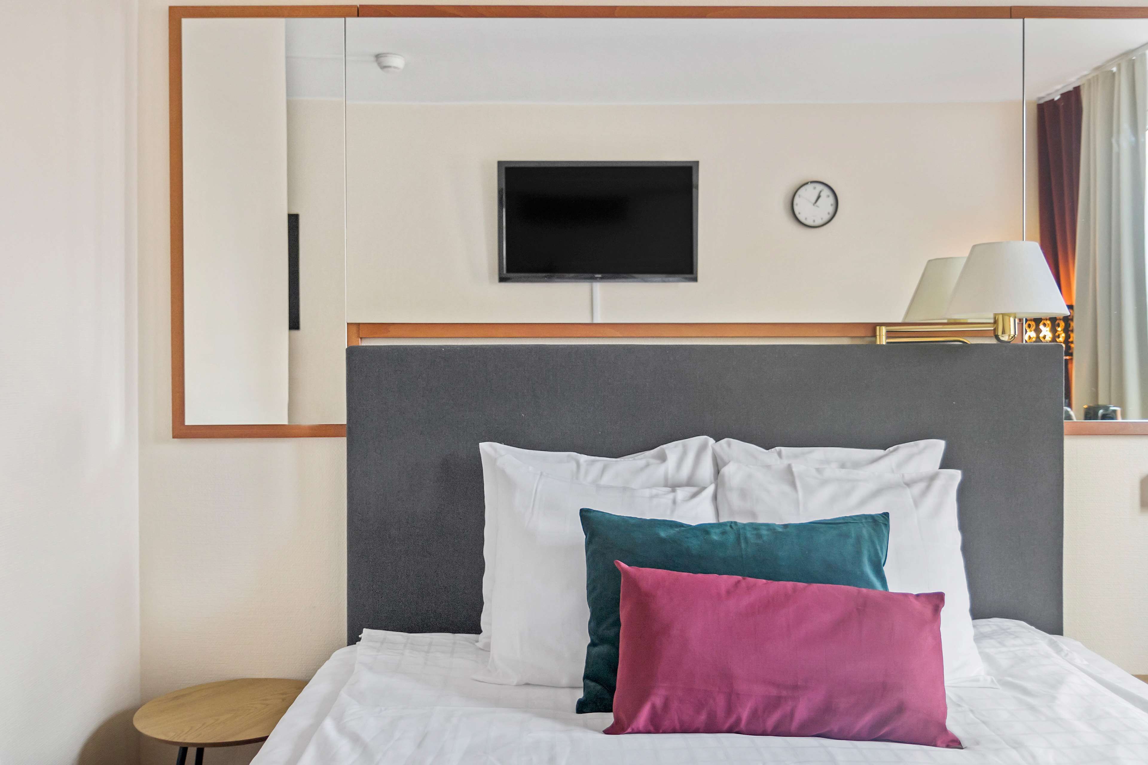 A hotel room with a bed, a television mounted on the wall, a clock, and a lamp.