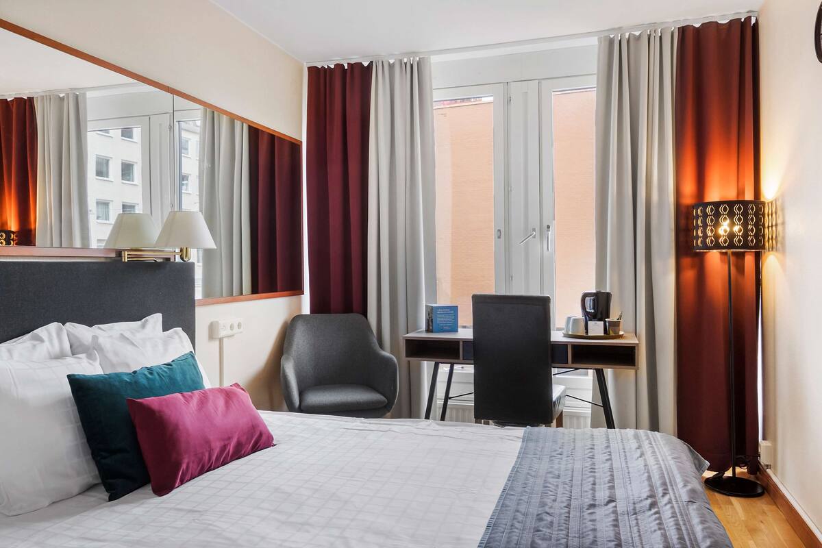 Une chambre d’hôtel avec un lit, deux chaises, un bureau, une lampe et une vue sur la ville grâce à de grandes fenêtres.