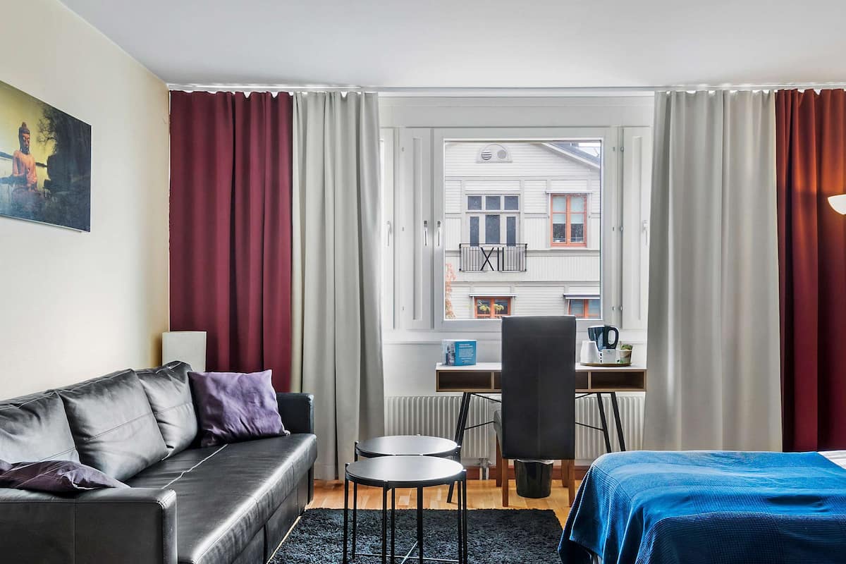 Une chambre d’hôtel avec un canapé en cuir noir, une couvre-lit bleue, une petite table ronde et une chaise. Il y a une fenêtre donnant sur un bâtiment à l’extérieur.