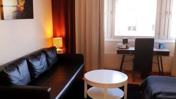 A modern living room with a black leather sofa, a white round table, and a desk with a chair. A painting is on the wall, and there is a window with curtains.