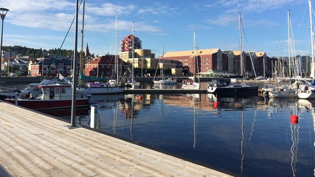 Uma marina com barcos atracados, um cais de madeira e edifícios ao fundo.