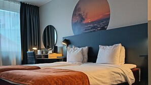Quarto de hotel com uma cama grande, uma escrivaninha com candeeiro, um espelho redondo e uma parede com uma pintura de pôr do sol.