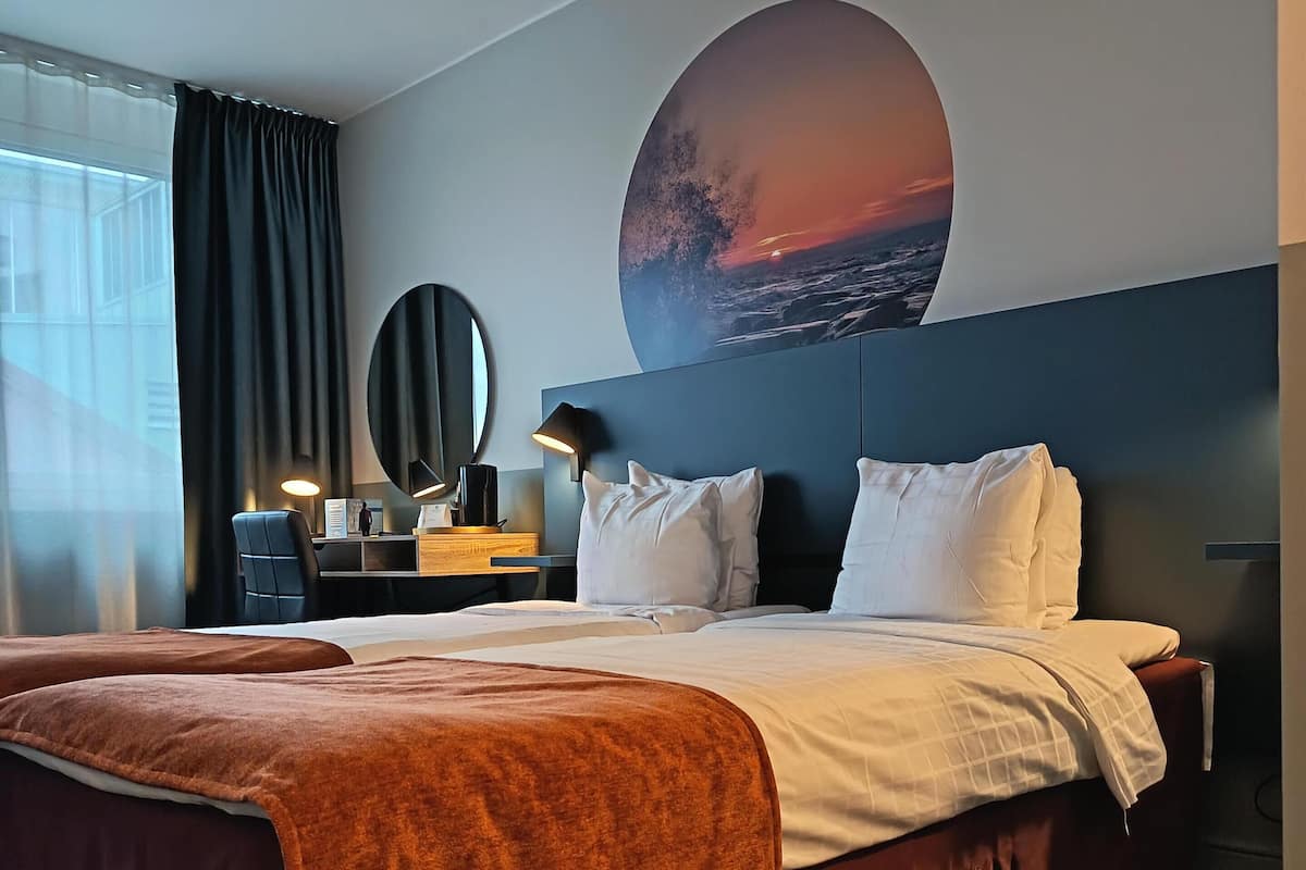 Une chambre d’hôtel avec un grand lit, un bureau avec une lampe, un miroir rond et un mur orné d’un tableau représentant un coucher de soleil.