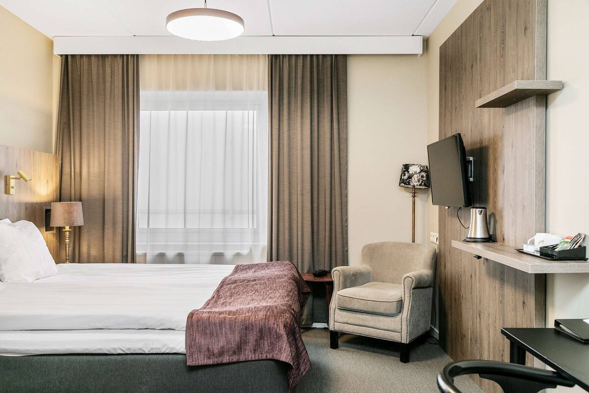 A hotel room with a bed, a chair, a desk, a television, and a window with curtains.