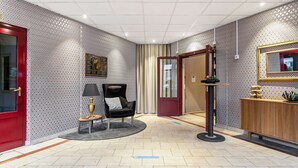 A hotel lobby with patterned wallpaper, a red door, a black chair, a small table with a lamp, a mirror, and a wooden cabinet. - Hotel Mälarporten (Vasteras)