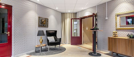 A hotel lobby with patterned wallpaper, a red door, a black chair, a small table with a lamp, a mirror, and a wooden cabinet.